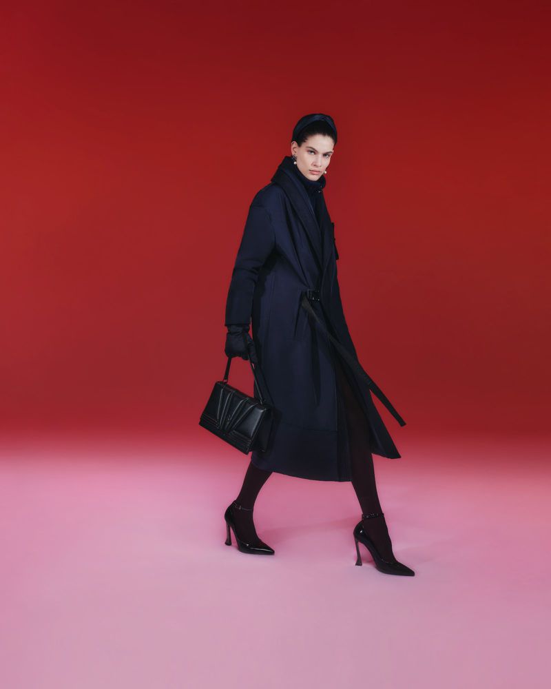 Model in a long navy belted coat and black stiletto heels carrying a black leather handbag against a red studio backdrop.