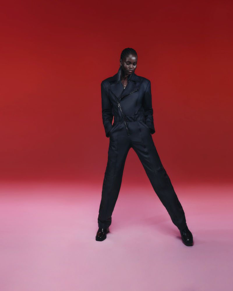 A tall, slender African American woman in a black suit stands in a studio against a red and pink backdrop in a fashion style photo.