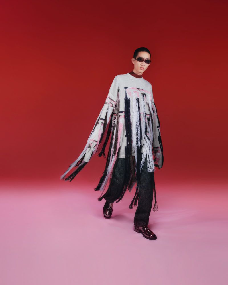 Model Benji Xu wearing a fringed patterned sweater and sunglasses against a gradient red studio background