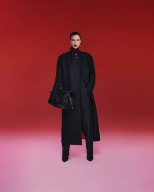 Model Irina Shayk wearing a dark coat and trousers while holding a black handbag against a red background