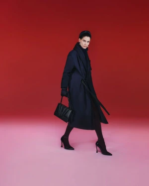 Model in a long navy belted coat and black stiletto heels carrying a black leather handbag against a red studio backdrop.