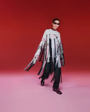 Model Benji Xu wearing a fringed patterned sweater and sunglasses against a gradient red studio background