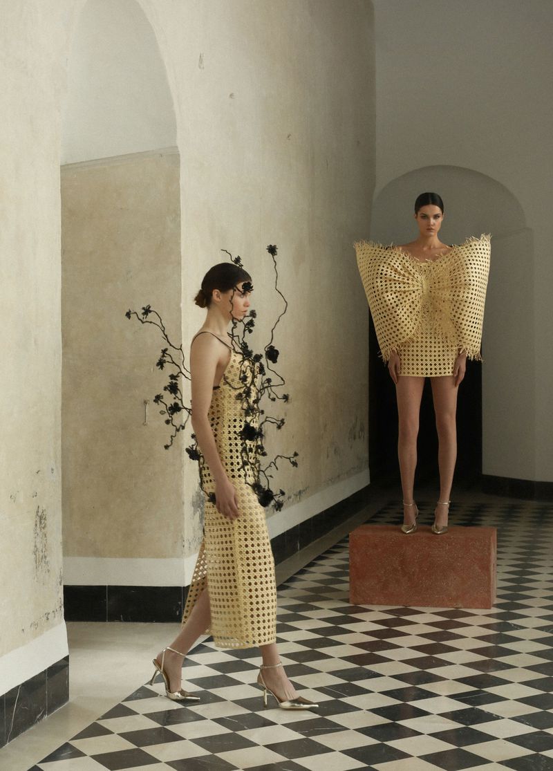 Two women pose in woven yellow dresses in an arcade. One woman walks toward the viewer, while the other stands on a stone block.