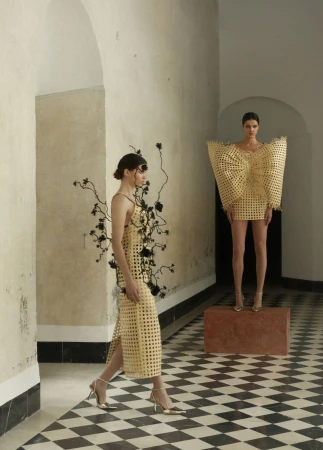 Two women pose in woven yellow dresses in an arcade. One woman walks toward the viewer, while the other stands on a stone block.