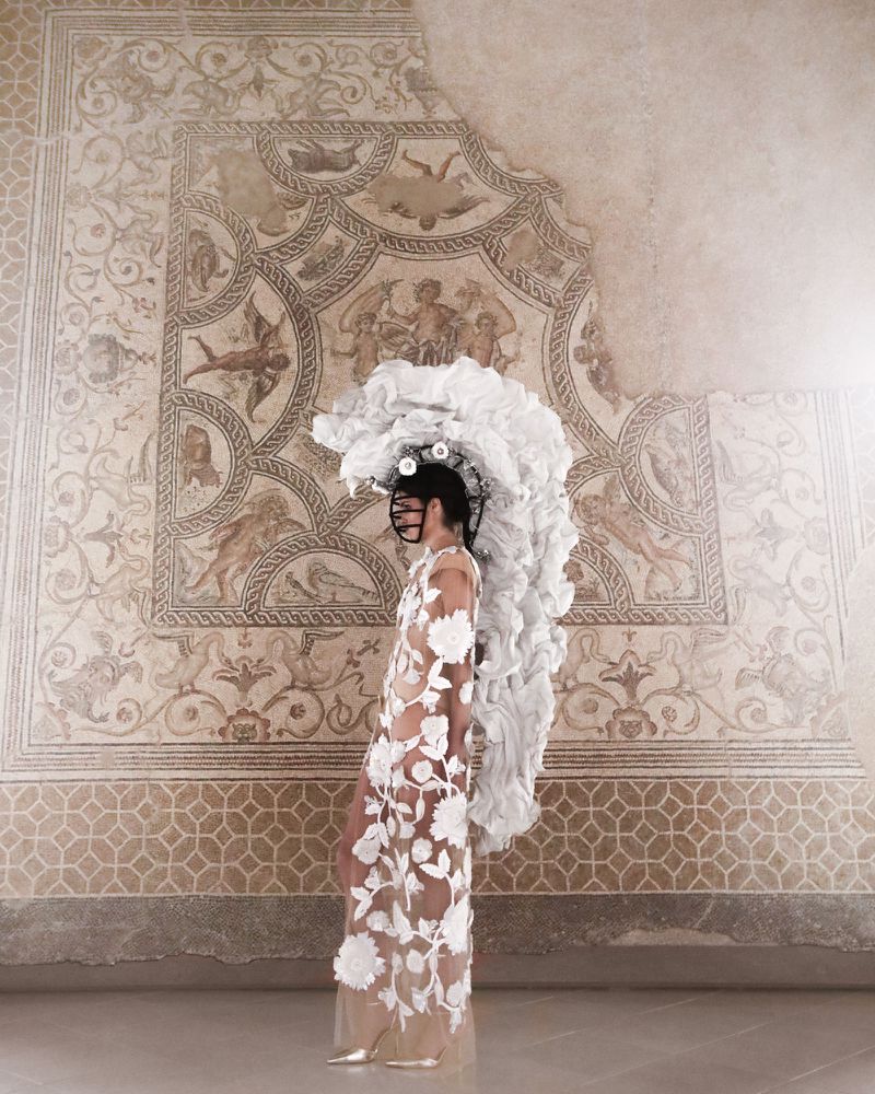 Fashion model poses in a sheer floral gown with a large ruffled headpiece against a cracked mosaic wall in an indoor setting.