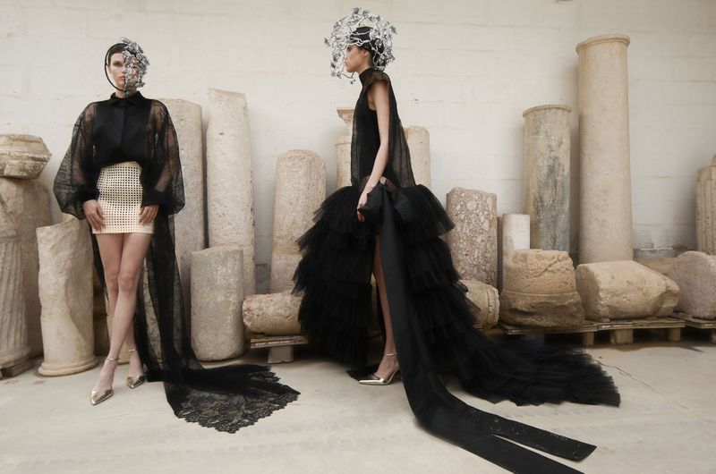 Two fashion models in black gowns and ornate headpieces pose among ancient stone columns, creating a dramatic and artistic composition.