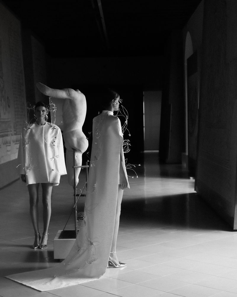 Black and white fashion shot features two women in white dresses and heels, posed in a hallway next to a headless statue.