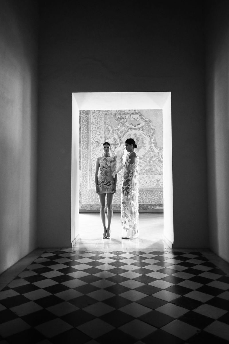 Black and white fashion shot of two women in embroidered dresses standing in a hallway with a black and white checkered floor.
