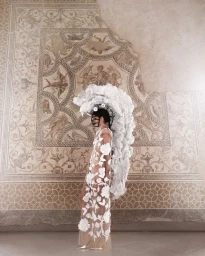Model Maria Parr wearing white couture gown and elaborate feathered headdress against ancient mosaic wall