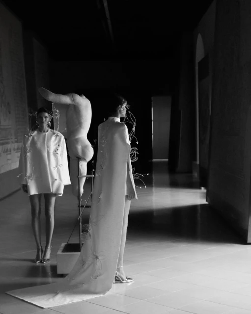 Two female models in white haute couture dresses posing inside a gallery beside a sculptural art installation.
