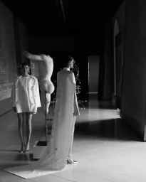 Two female models in white haute couture dresses posing inside a gallery beside a sculptural art installation.
