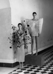 Two female models in avant-garde white cutout dresses with black branch and bow motifs standing on checkered floor.
