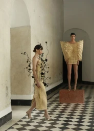Two models in structured woven dresses walking in a high-ceilinged hallway with black-and-white checkered floors and arched plaster walls.
