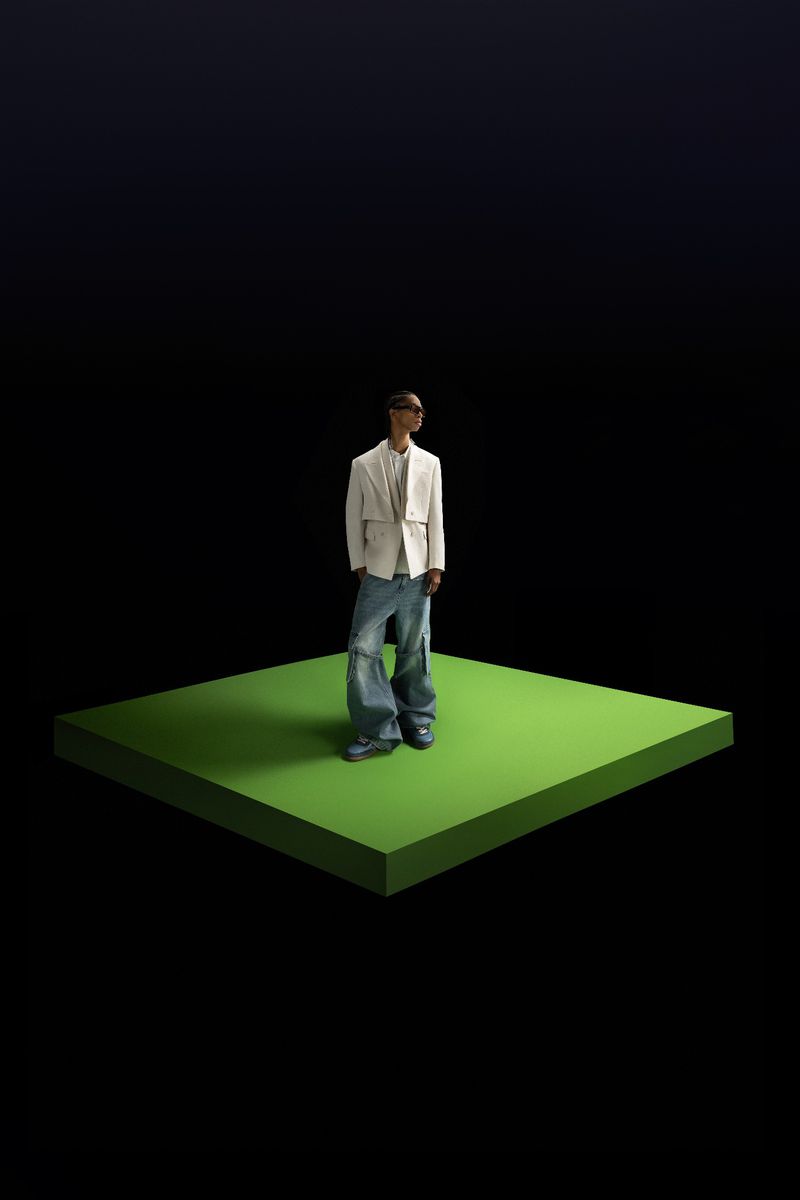 Full-length shot of a person in a white jacket and jeans standing on a green platform against a dark background.