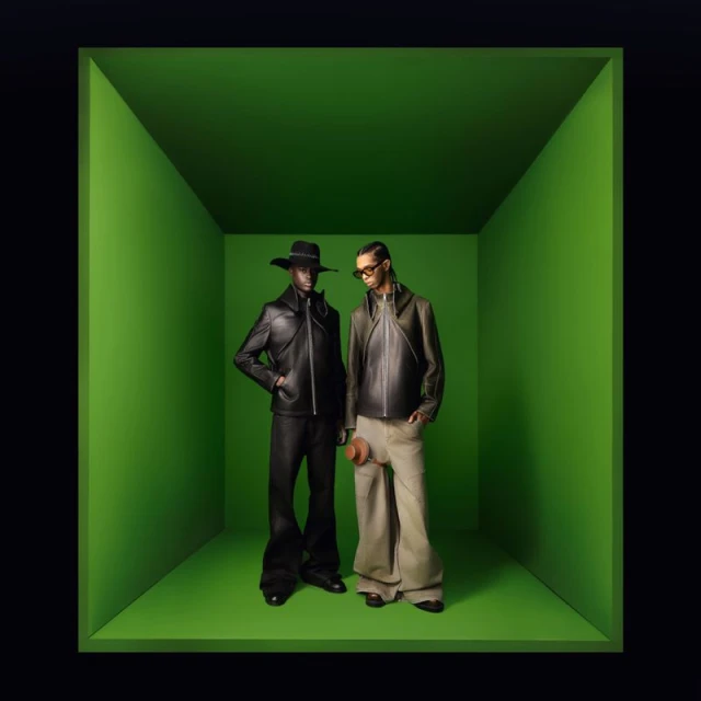 Two adult male fashion models pose in a bright green cubical photo studio wearing trendy designer leather jackets and flared pants.