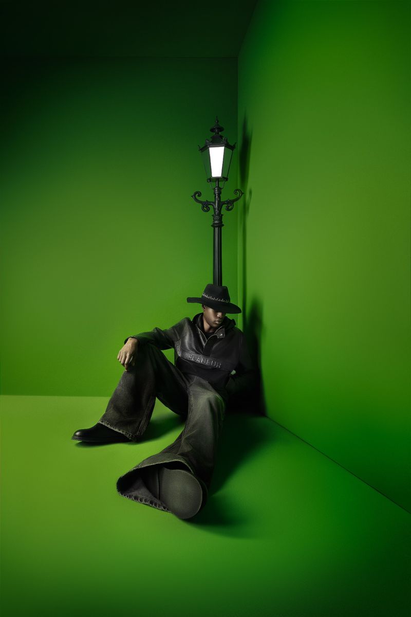 Adult male in hat and loose pants sits on a bright green floor next to a vintage-style black lamppost in the corner of a green room.
