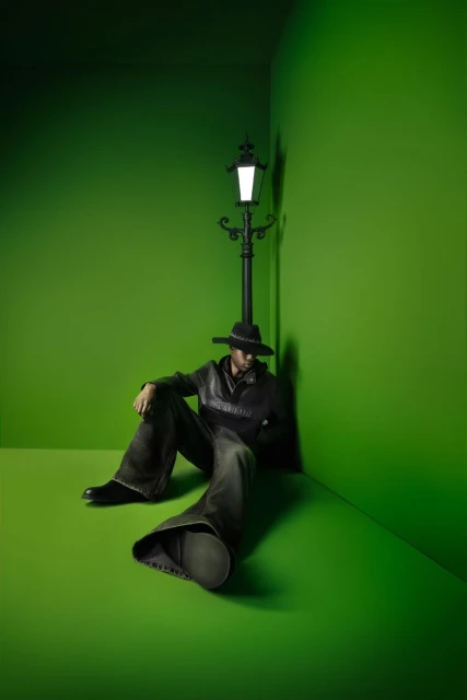 Adult male in hat and loose pants sits on a bright green floor next to a vintage-style black lamppost in the corner of a green room.