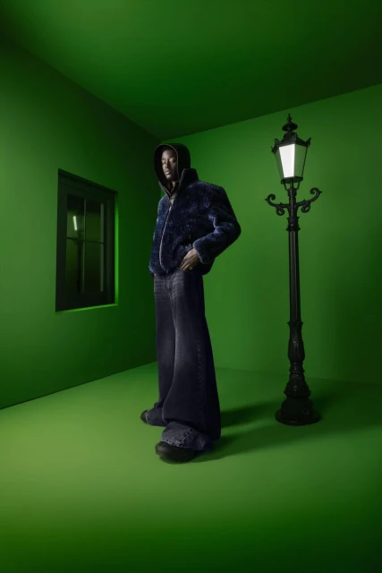 Full-length shot of adult male model in hooded jacket and wide-leg jeans, posing in a green room with a black window and lamp post.
