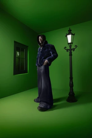 Full-length shot of adult male model in hooded jacket and wide-leg jeans, posing in a green room with a black window and lamp post.