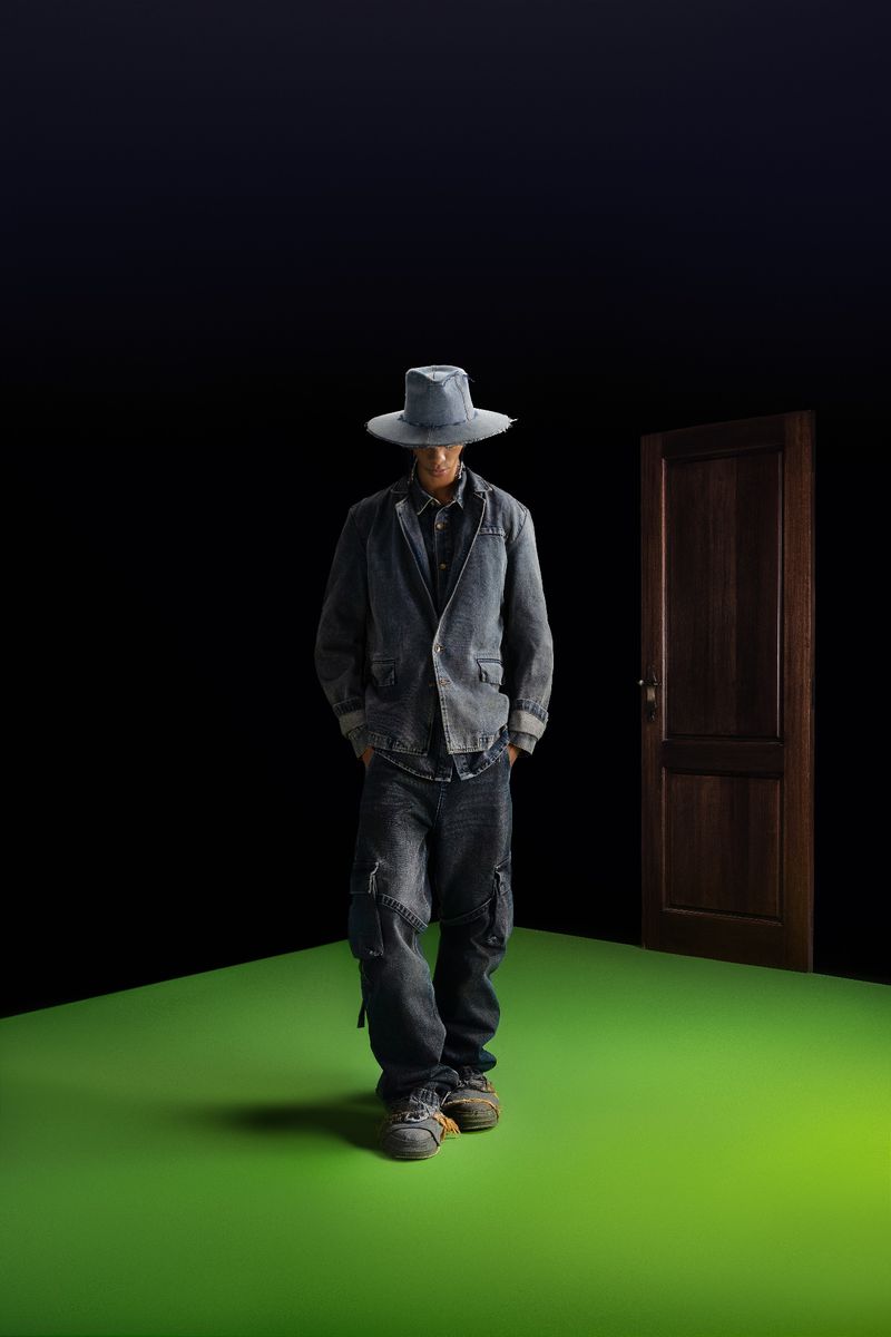 Model wearing a denim jacket, matching jeans, and a wide-brim hat standing on a bright green floor against a dark background with a wooden door.