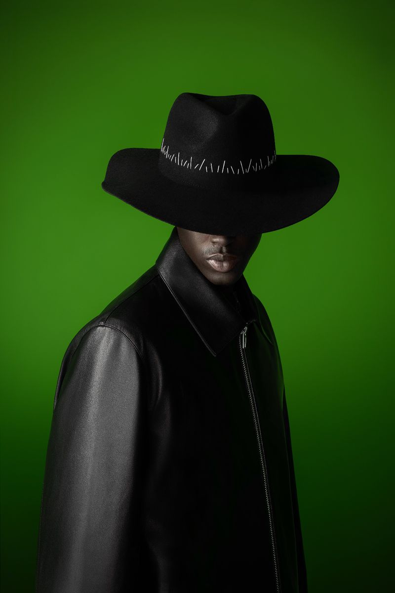 Portrait of an adult man wearing a black leather coat and a wide-brimmed hat against a green backdrop in a studio setting.