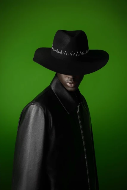 Portrait of an adult man wearing a black leather coat and a wide-brimmed hat against a green backdrop in a studio setting.