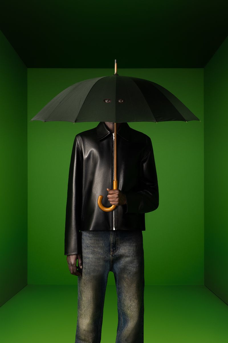 Model in a black leather jacket and jeans holding a black umbrella with cut-out eyeholes in a green studio room.