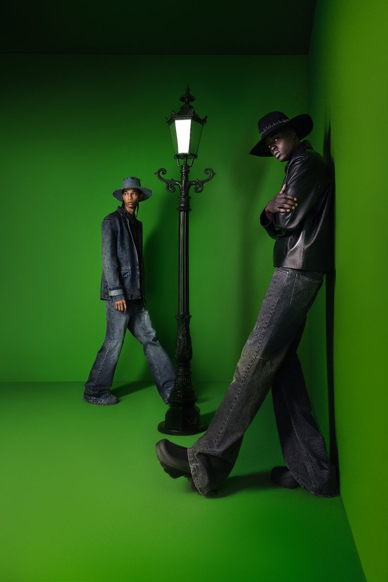 Two men pose in a green studio with a vintage streetlamp. One in a leather jacket leans against the wall. Fashion editorial shot.