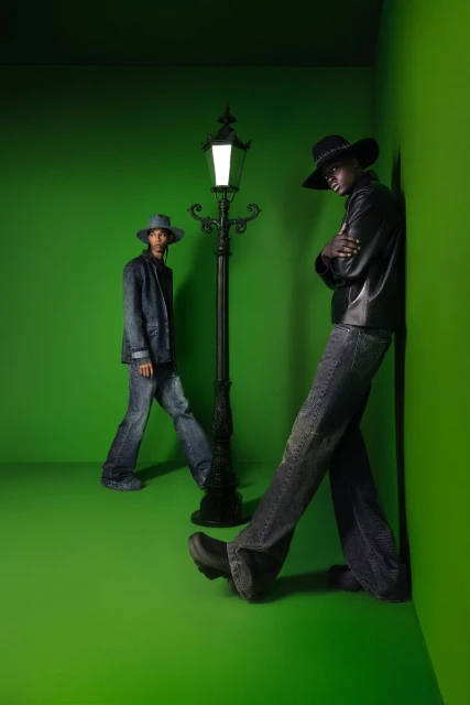 Two men pose in a green studio with a vintage streetlamp. One in a leather jacket leans against the wall. Fashion editorial shot.