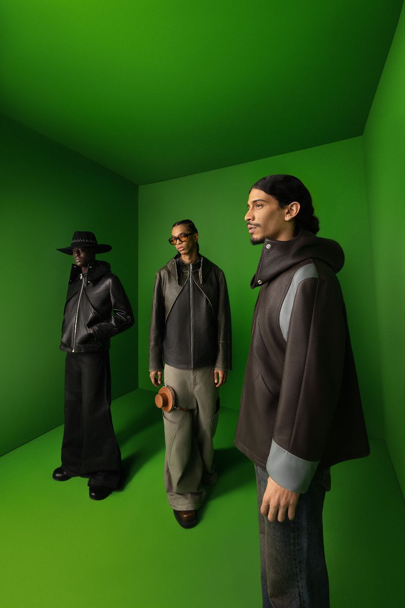 Three men model dark colored jackets in a simple green studio. One wears jeans, another loose pants, one wears a long dress-like skirt.