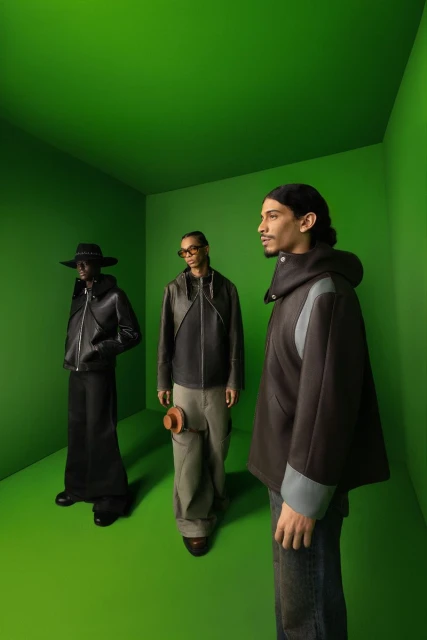 Three men model dark colored jackets in a simple green studio. One wears jeans, another loose pants, one wears a long dress-like skirt.