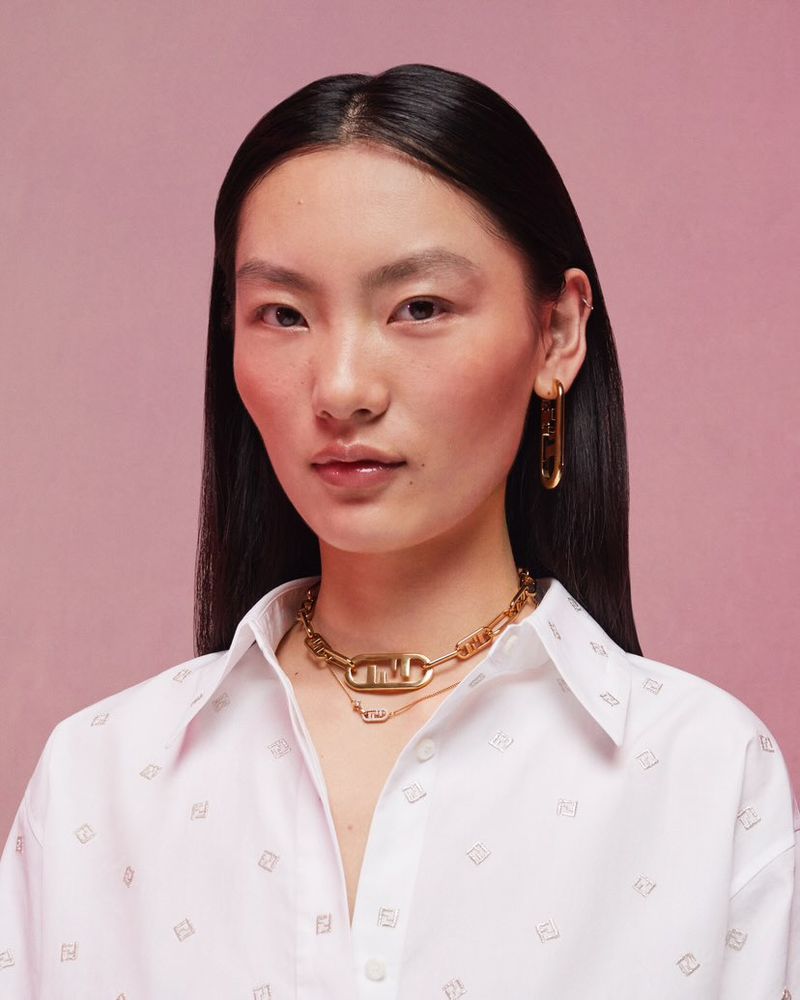 Model wearing Fendi O'Lock gold jewelry and a white patterned shirt against a pink seamless background.