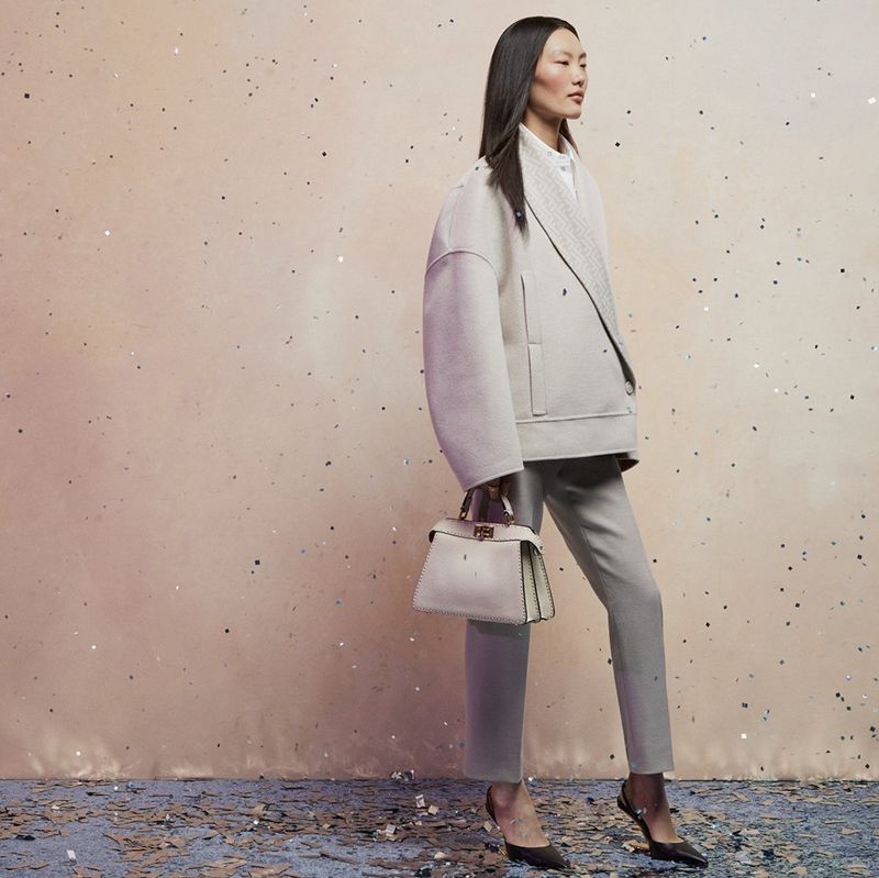 Elegant Asian woman showcases a luxury handbag and wears a cream-colored oversized jacket with matching pants in a fashion studio shot.