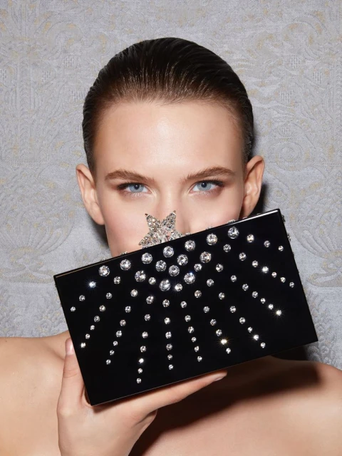 Woman with fair skin and slicked-back hair holds a black clutch bag adorned with crystals in front of her face in a beauty shot.