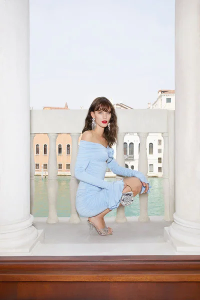 Model Coline Leclere crouching in a light blue off-the-shoulder mini dress and embellished heels on a stone balustrade balcony.