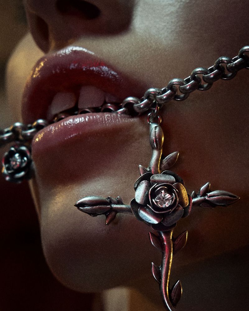 Close-up of a person's mouth with glossy red lipstick, biting on a silver chain necklace with a rose-shaped cross pendant.