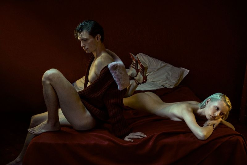 Male and female models pose intimately on a red satin bed in a moody, dark lit studio, capturing a raw and sensual moment.