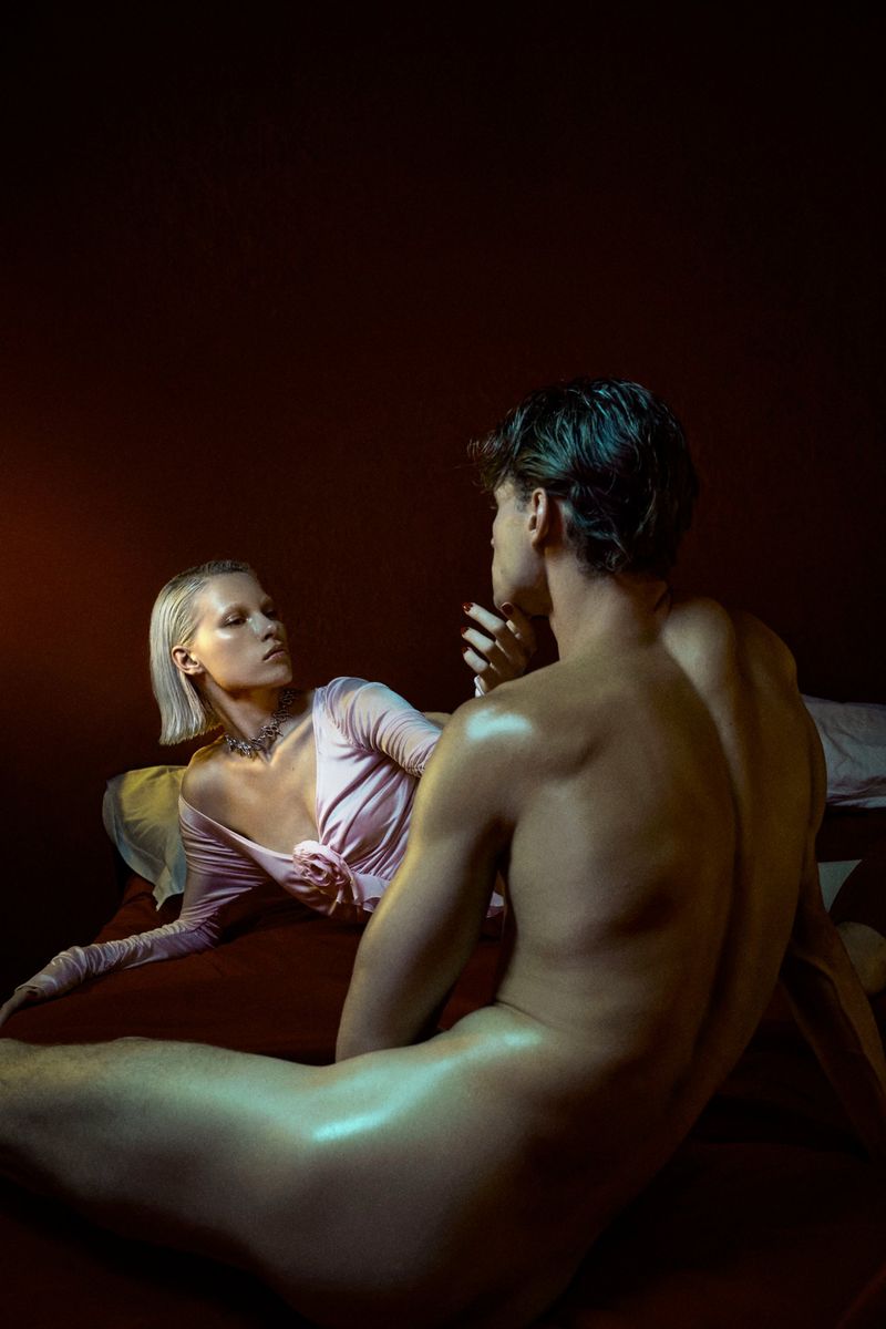 Romantic studio shot of shirtless man facing woman with pale skin in pink top on bed, set against dark red background.