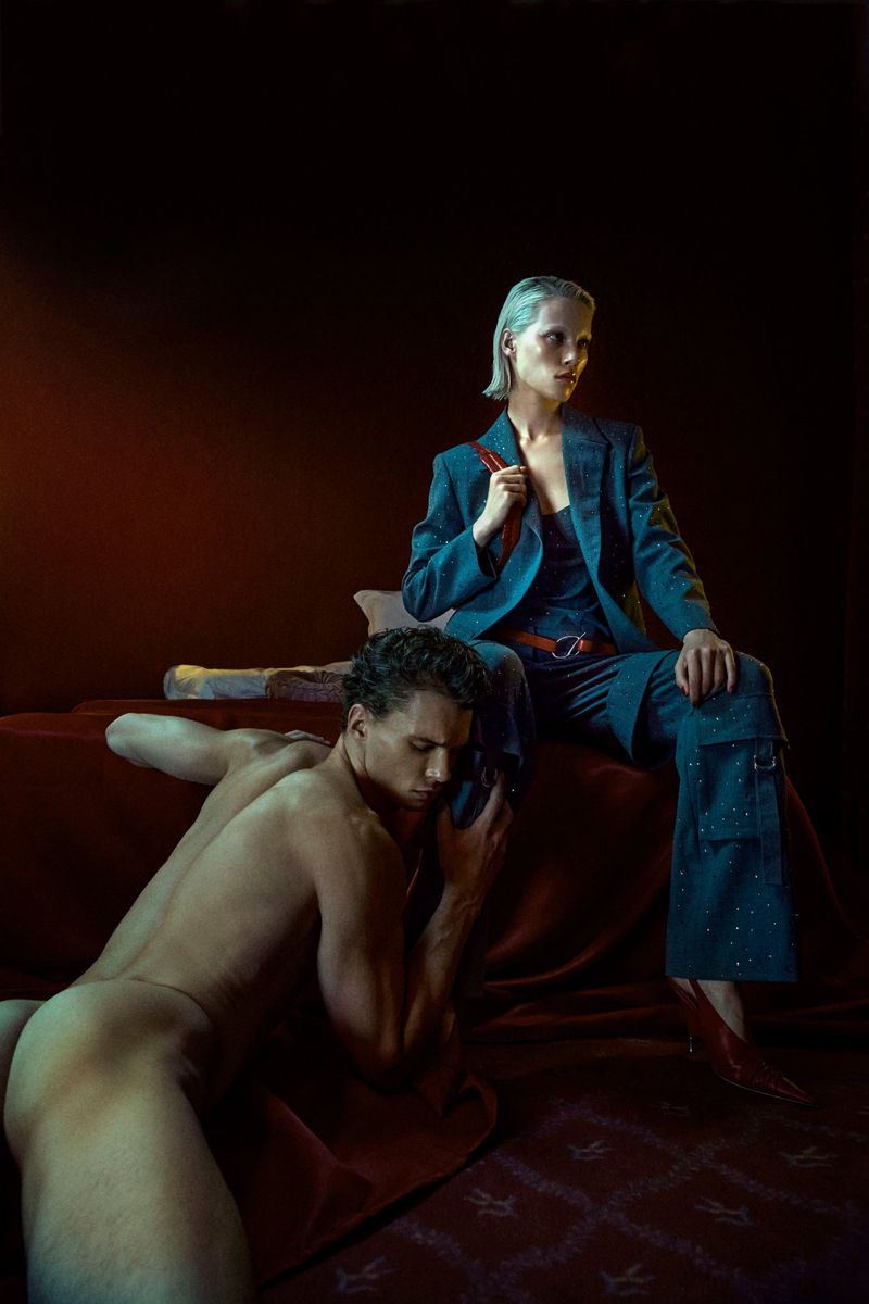 Striking portrait of a woman in a suit seated with a nude man at her feet, captured in rich reds and cool blues in a studio setting.