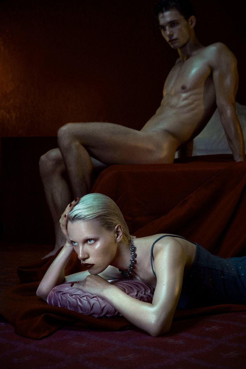 Two adult models in a photo studio. A shirtless man sits in the background while a platinum blonde woman reclines on a bed.