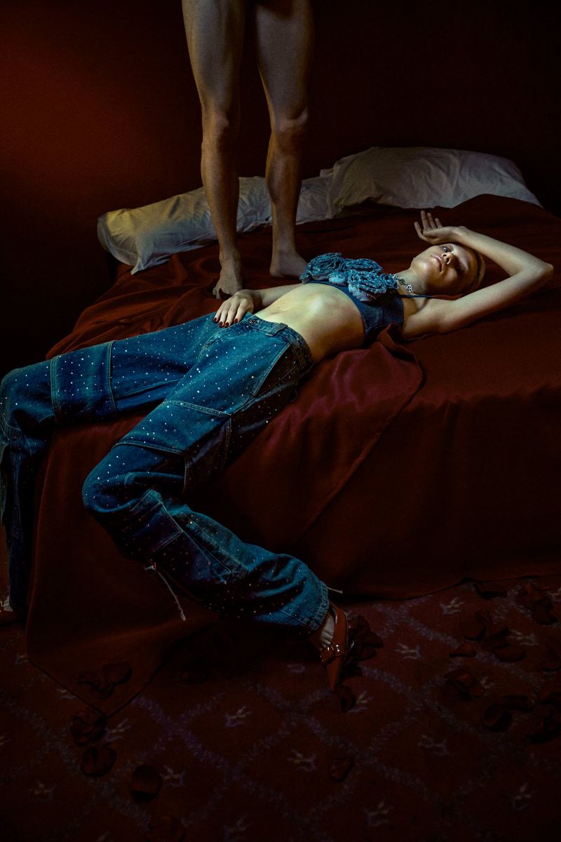 Blonde fashion model in bejeweled denim bra and pants reclines on a red velvet bed as a man stands at the head of the bed.