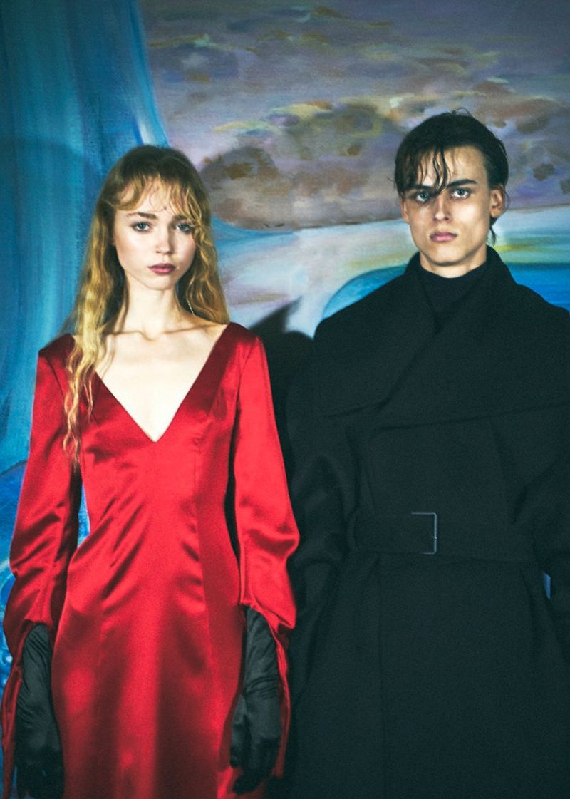 A fair-skinned young couple stands together in a studio setting. The woman is in a red satin dress, and the man is in a black overcoat.