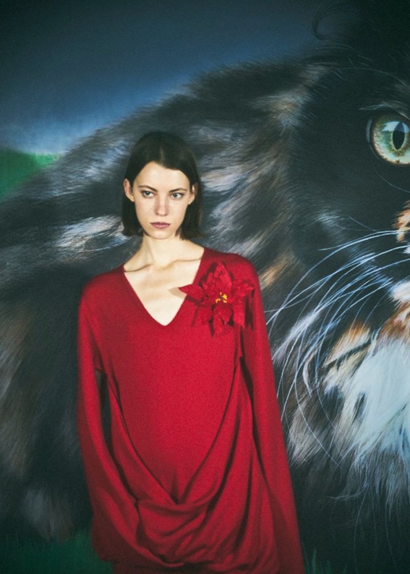 A woman in a red dress stands in front of a backdrop with a large cat face in a medium studio shot, illuminated with bright lighting.