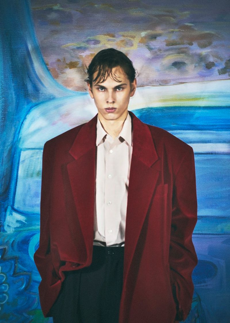 Model wearing an oversized burgundy blazer and pale button-down shirt against a blue painted scenic backdrop.