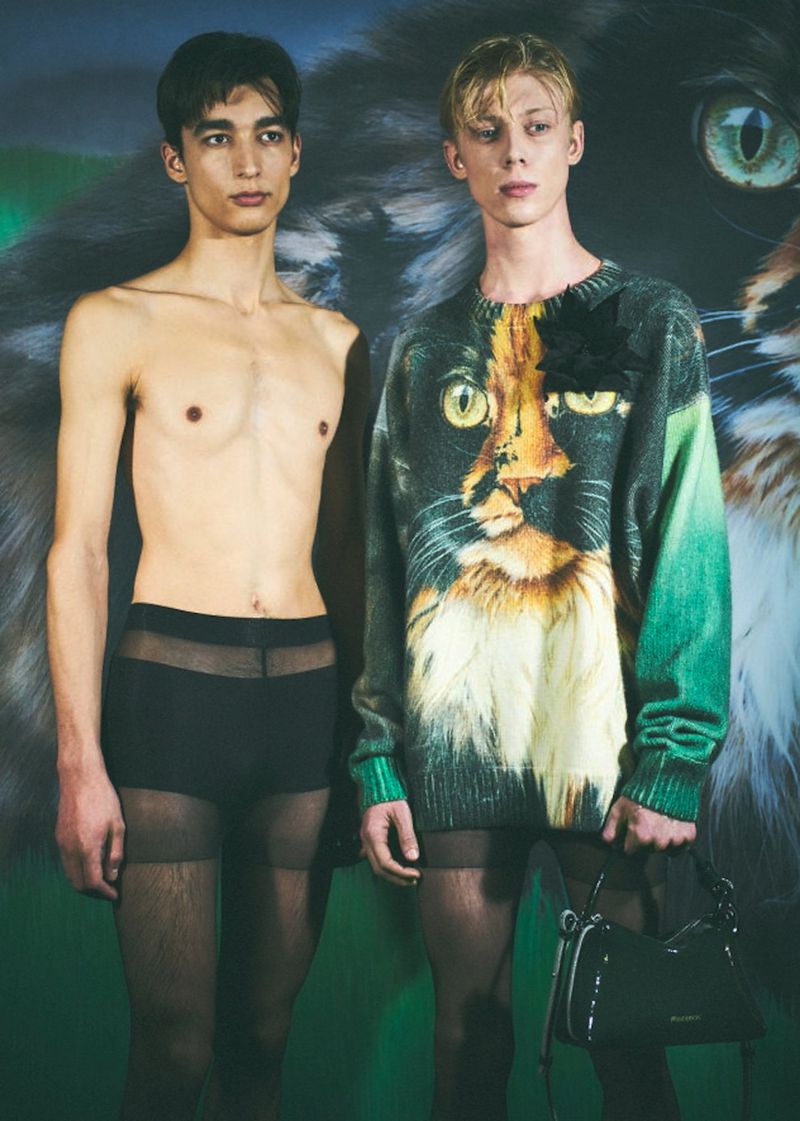 Two men pose in front of a cat backdrop. One is shirtless wearing sheer black shorts with tights, and the other wears a cat sweater.