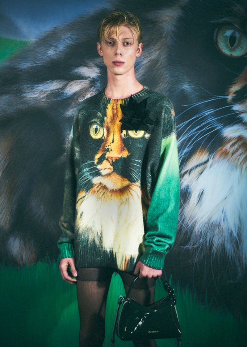 A young adult poses in a fashion shot, wearing a cat-themed sweater, accessorized with a black flower, and a patent black handbag.
