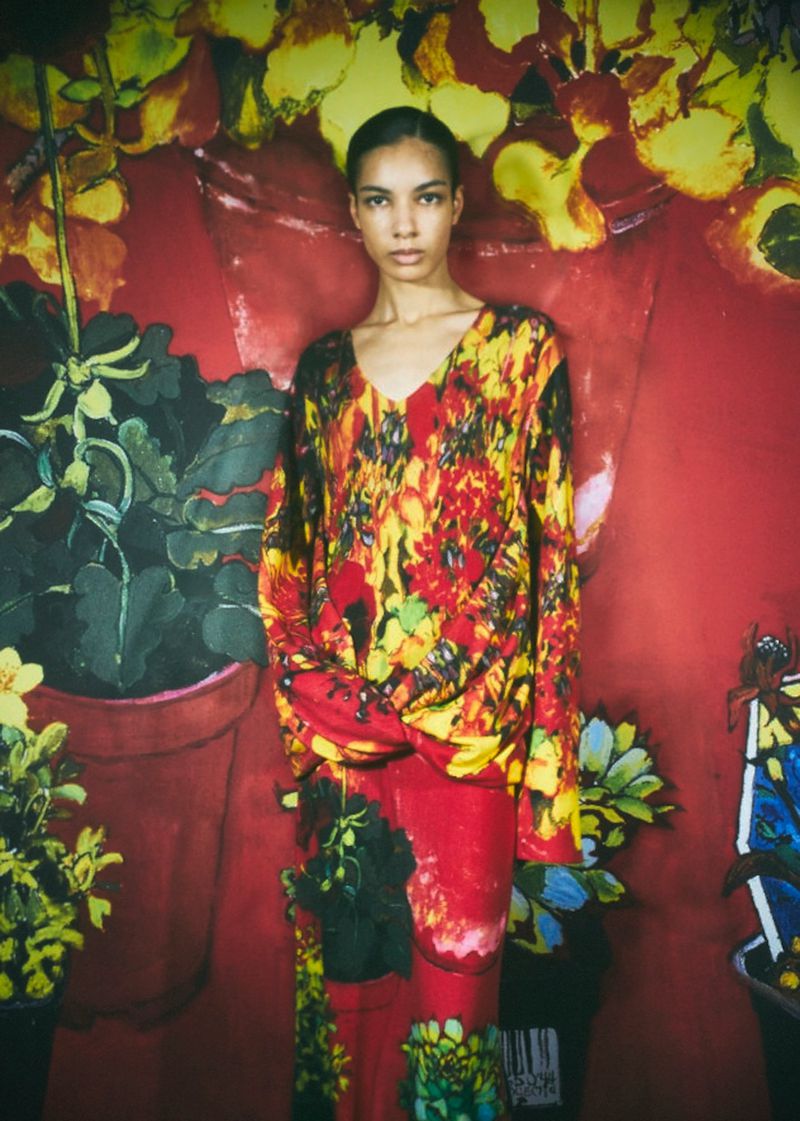 Fashionable young woman poses in red patterned outfit in front of floral backdrop in a studio, showcasing unique style and composition.