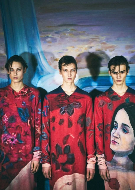 Three young adult men wearing patterned long-sleeved shirts with a painted background of a waterfall and a partial painting of a woman.