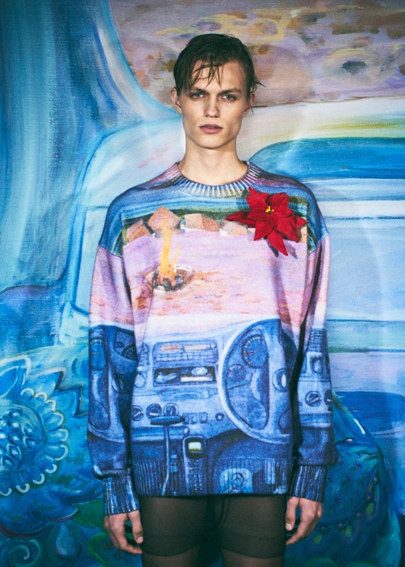 Male model wearing a graphic-print sweater poses against a painted blue backdrop. The image features a red flower accent.
