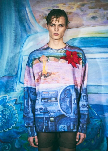 Male model wearing a graphic-print sweater poses against a painted blue backdrop. The image features a red flower accent.
